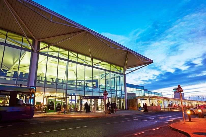 Bristol Airport terminal building with a plane taking off in the background, with a caption 'Bristol Airport Removes Advert After Public Backlash'