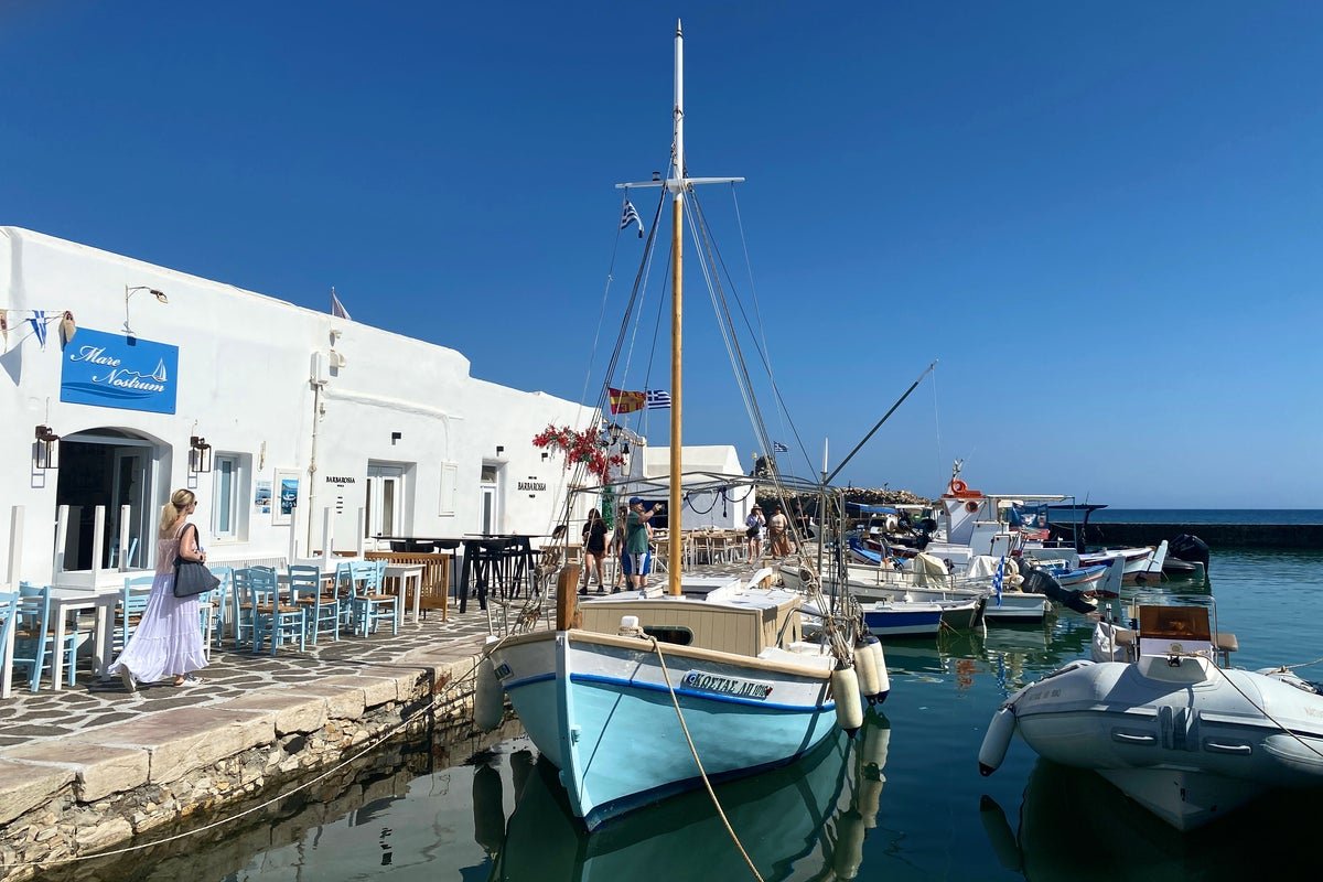 A passport control officer stamps a UK traveller's passport at a Greek airport, with a backdrop of Greek flags and a sign reading 'Welcome to Greece'. The image captures the moment when a UK traveller enters Greece, highlighting the country's unique approach to border control and its decision to ditch the EU's entry-exit biometrics system for British travellers. The colour of the Greek flag and the vibrant atmosphere of the airport reflect the country's rich culture and history, making it an attractive destination for tourists from around the world.