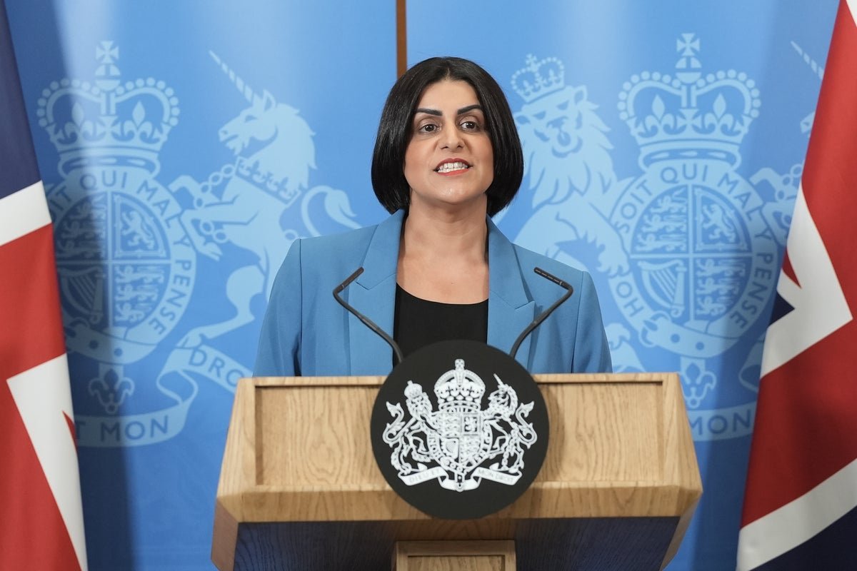Shabana Mahmood standing confidently in front of a UK flag, with a mixture of people in the background, some of whom are shouting, while she engages with them, with a description of the scene, including the colour and atmosphere of the moment, highlighting the primary keyword, Reform UK