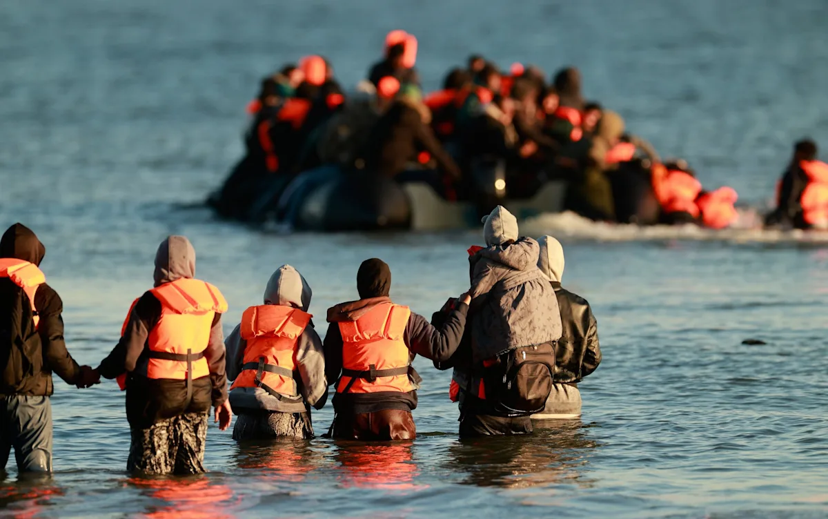 A group of migrants walking along a road, with a French flag visible in the background, symbolising their journey from Belgium to Britain with French assistance, highlighting the complex migration pattern and the role of France in this phenomenon