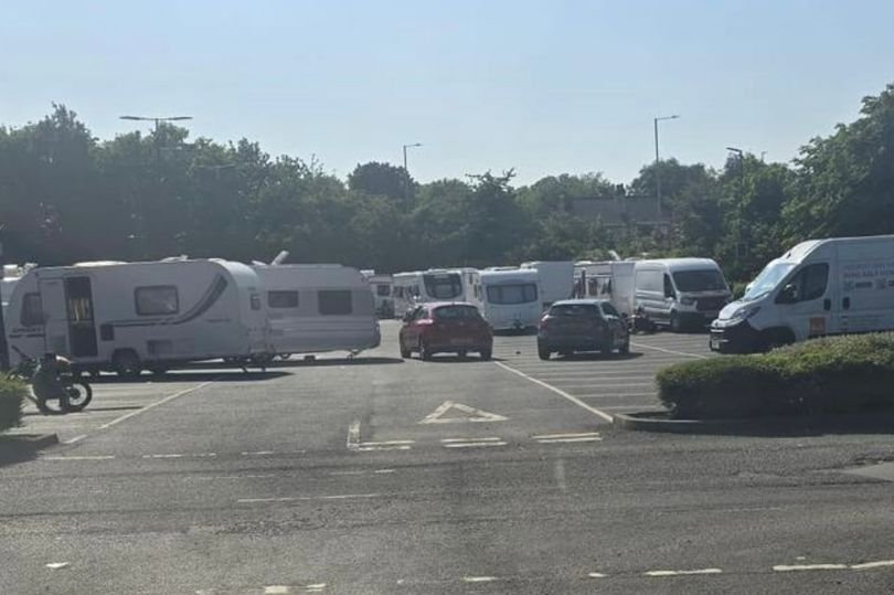Traveller campsite near B&Q and IKEA in Wednesbury, with caravans and vehicles parked on a gravel plot, surrounded by trees and a fence, highlighting the issue of unauthorised campsites and the need for designated traveller facilities