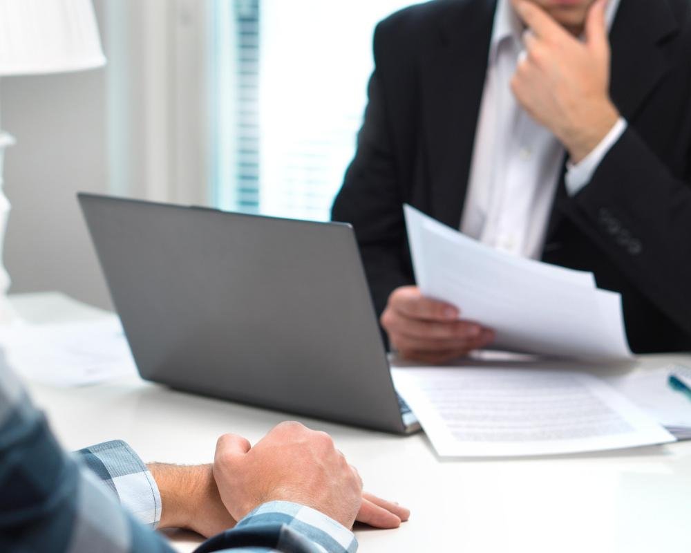 A recruiter sitting at a desk with a concerned expression, surrounded by stacks of unpaid bills and financial documents, with a photo of the Las Vegas strip in the background, highlighting the contrast between the company's financial woes and the lavish trip offered to staff