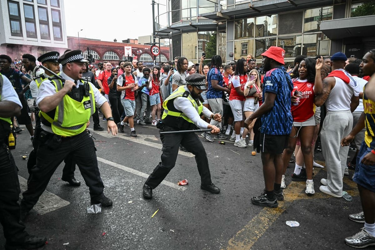 A crowd of Arsenal fans celebrating in the streets, with a flare being set off in the background, sparking a hotel fire and causing chaos and destruction, highlighting the need for safety and security at public events, with the primary keyword being football fandom.