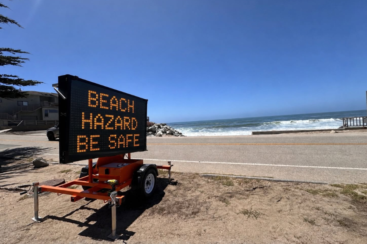 A dramatic image of a wave crashing on a California beach, with a warning sign in the foreground, highlighting the dangers of sleeping on the beach and the importance of beach safety and awareness of coastal hazards