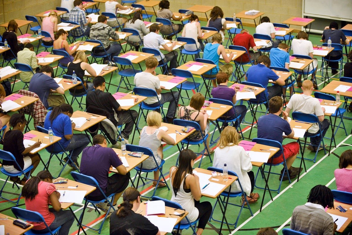 A group of students protesting outside a school, holding signs that read 'Fair Exams Now' and 'Review the A-Level Exams', with a caption that reads 'The A-level exams have sparked controversy, with many claiming they were more challenging than previous years'
