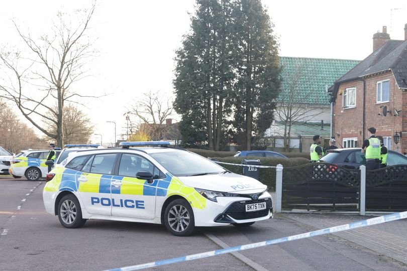 A West Midlands Police car parked outside a station, with a detective's badge and flowers in the foreground, symbolising the tragic loss of a police detective who died from burn injuries in the back of a parked car, with the primary keyword 'West Midlands Police detective' naturally included