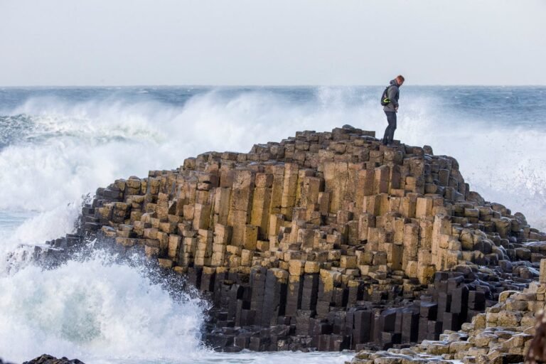 The Giant's Causeway in Northern Ireland, featuring distinctive basalt columns and a unique rock formation, with the sea crashing against the shore in the background, under a colourful sky with a mix of blue and grey hues, with a few visitors walking along the coastline, taking in the breathtaking views of this incredible natural wonder