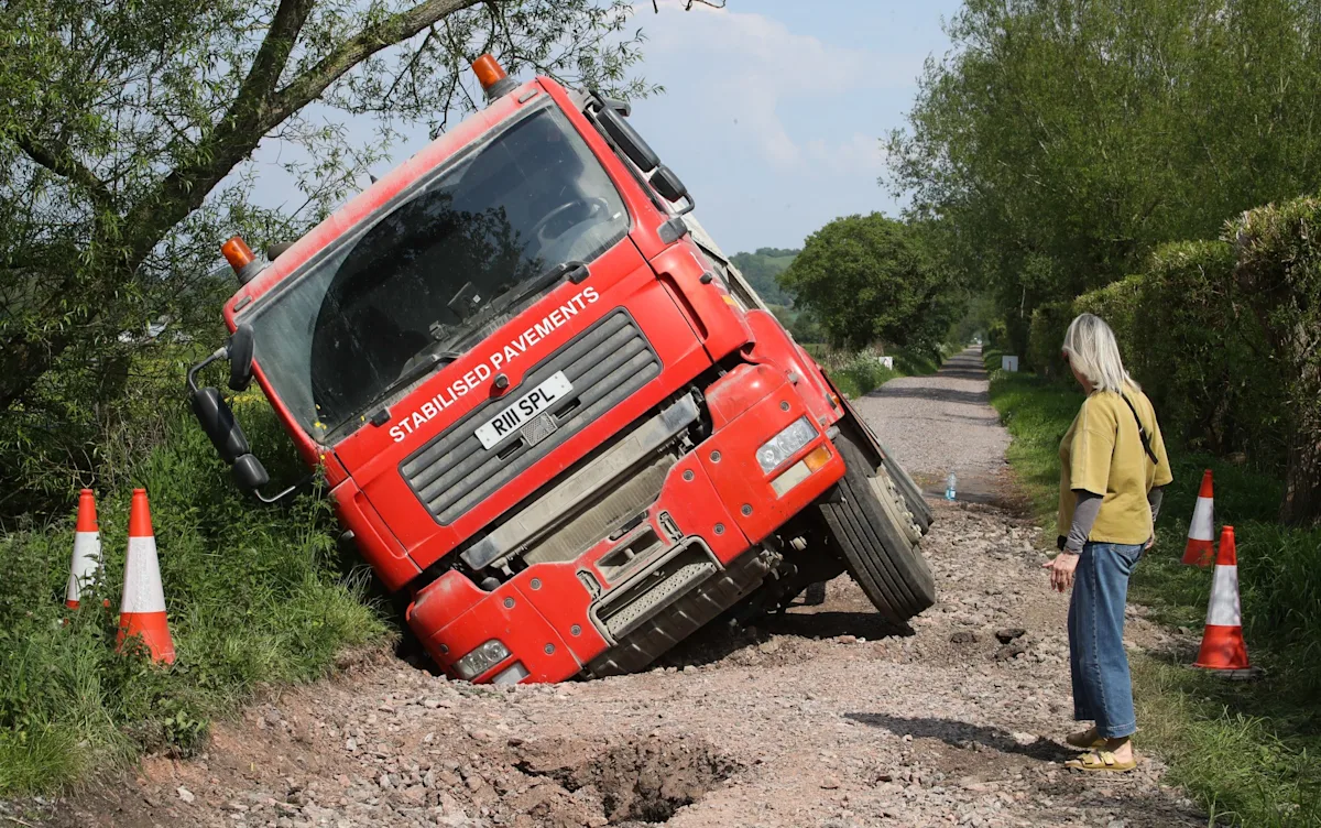 A pothole repair truck stuck in a deep pothole on a busy road, with a crew of workers trying to rescue it, highlighting the challenges of pothole repair and the need for better road maintenance