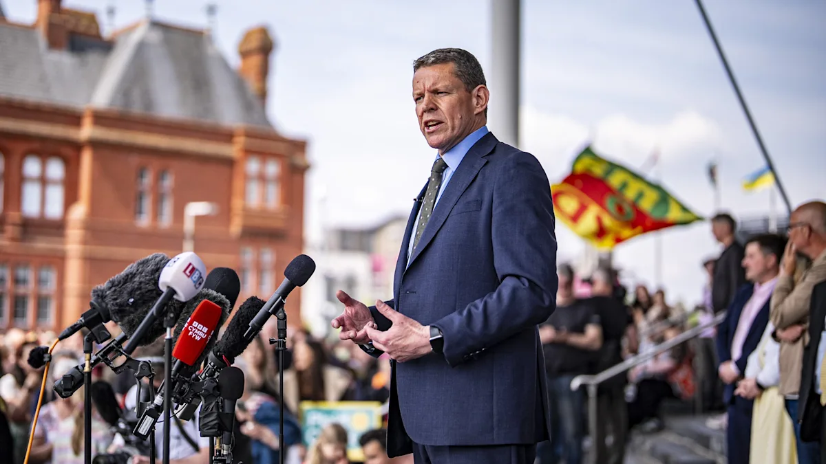 Rhun ap Iorwerth, the new First Minister of Wales, smiling and shaking hands with a colleague, with the Welsh flag in the background, symbolising his new role and responsibilities in Welsh politics