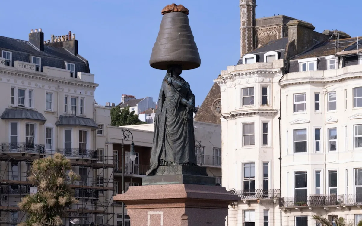 A statue of Queen Victoria with a Walnut Whip on her head, sparking a mixture of reactions from the public, with the colourful treat standing out against the more subdued tones of the statue, raising questions about the cultural significance of the Walnut Whip