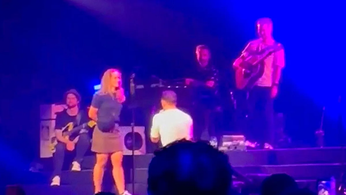 A man proposing to his girlfriend on stage in front of a large crowd, with a mix of shocked and amused expressions on the faces of the audience members, highlighting the unpredictability of public proposals and the importance of being mindful of the other person's boundaries and feelings