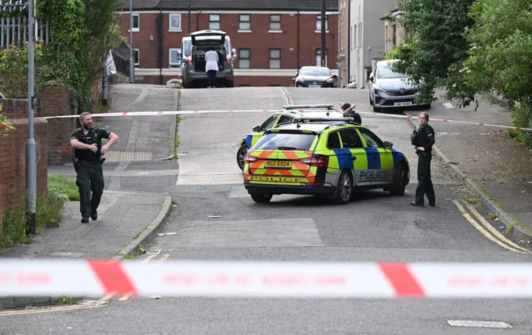 A photo of a police car in Belfast, with a Sudanese migrant in the background, highlighting the issue of attempted beheading and migrant behaviour in the UK